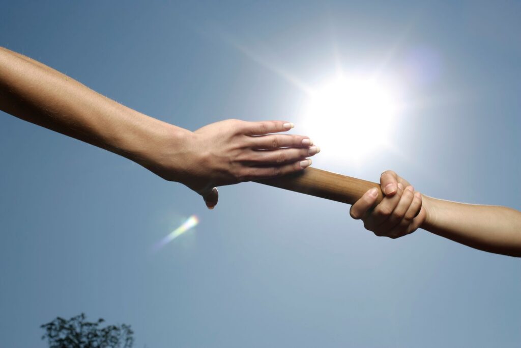 Two hands passing a baton under a bright sun, symbolizing teamwork and cooperation.